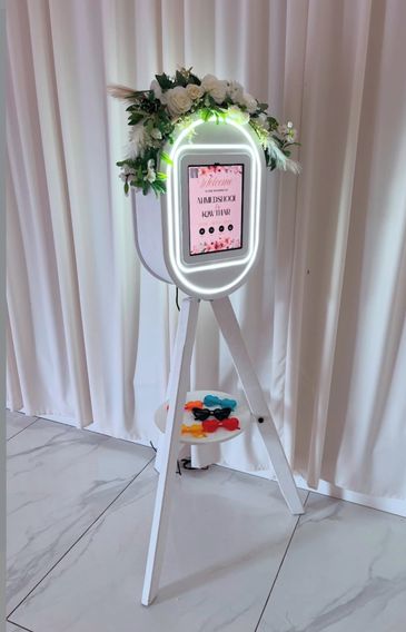 Elegant photo booth with floral decor and colorful props on a white tripod stand.