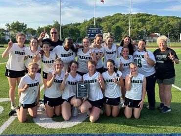 Girls' lacrosse team celebrating with a trophy on the field.