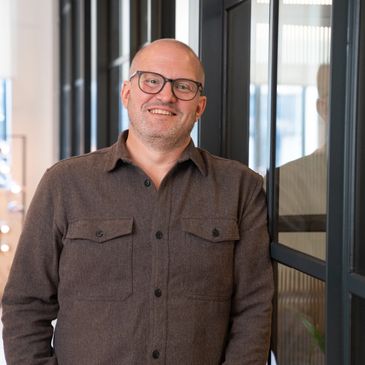Smiling man in glasses leaning against glass wall in a modern office.