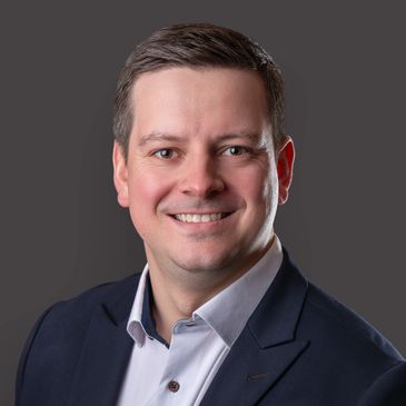Smiling man in a navy blazer and light blue shirt against a gray background.