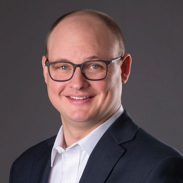Smiling man with glasses in a suit and white shirt against a gray background.