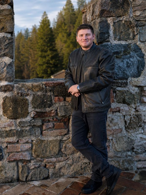 Man in black leather jacket standing by a stone wall.
