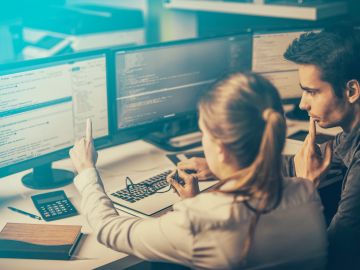 Two people collaborating on computer programming at dual monitors.