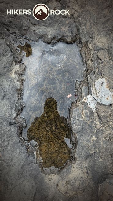 A man taking a picture of his reflection in a water puddle by a creek. 