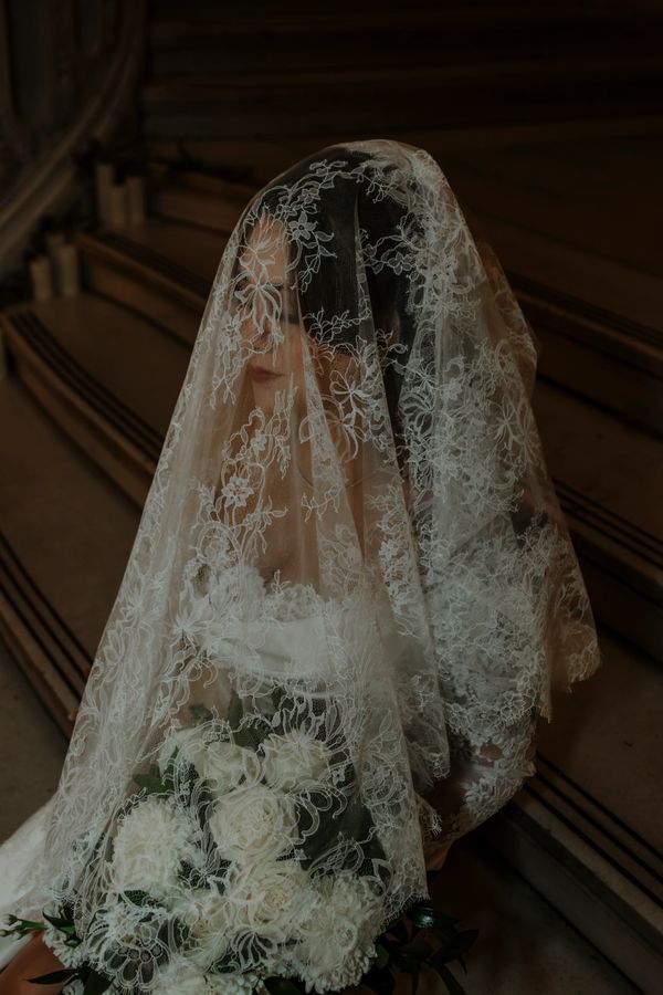 Bride with lace veil and white bouquet sitting on stairs.