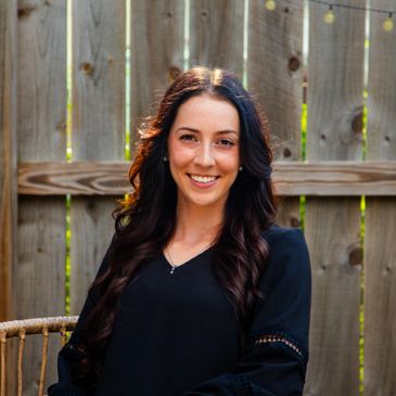 Smiling woman with long dark hair sitting outdoors against a wooden fence.