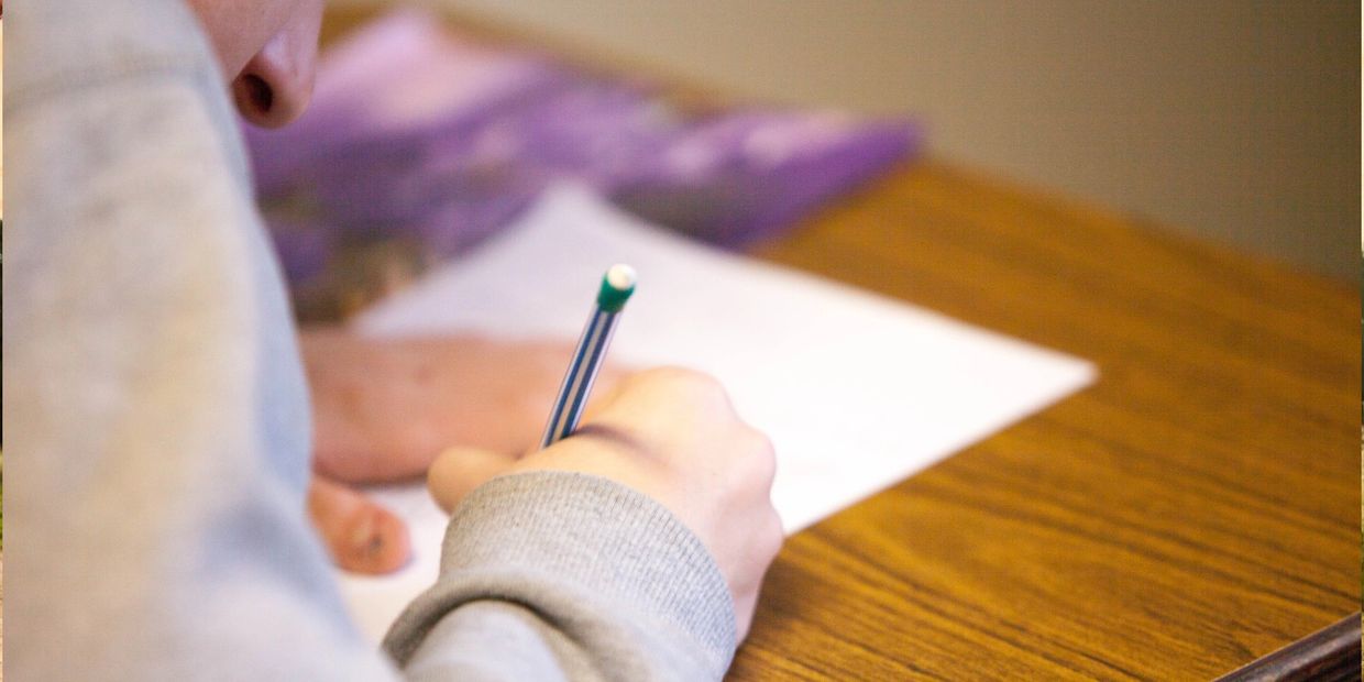 Student writing on paper with a pencil at a wooden desk.