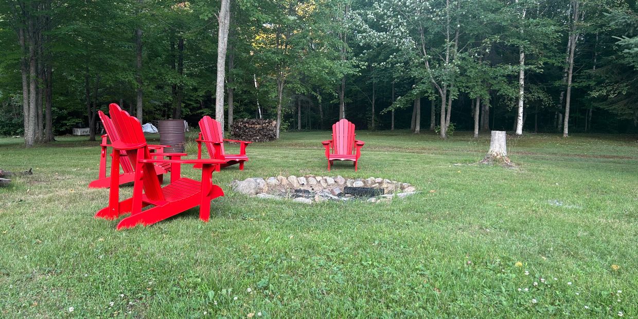 Red Adirondack chairs around a stone fire pit on a grassy lawn near trees.