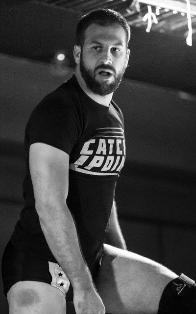 Drew Gulak posing during a professional wrestling match wearing a black Catchpoint Tee Shirt.