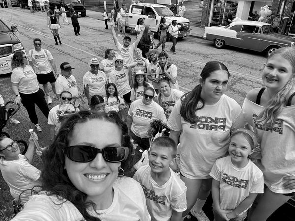 Group selfie at a colorful pride parade with matching shirts and rainbow face paint.