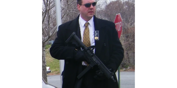Man in black coat and sunglasses holding a rifle near a stop sign.
