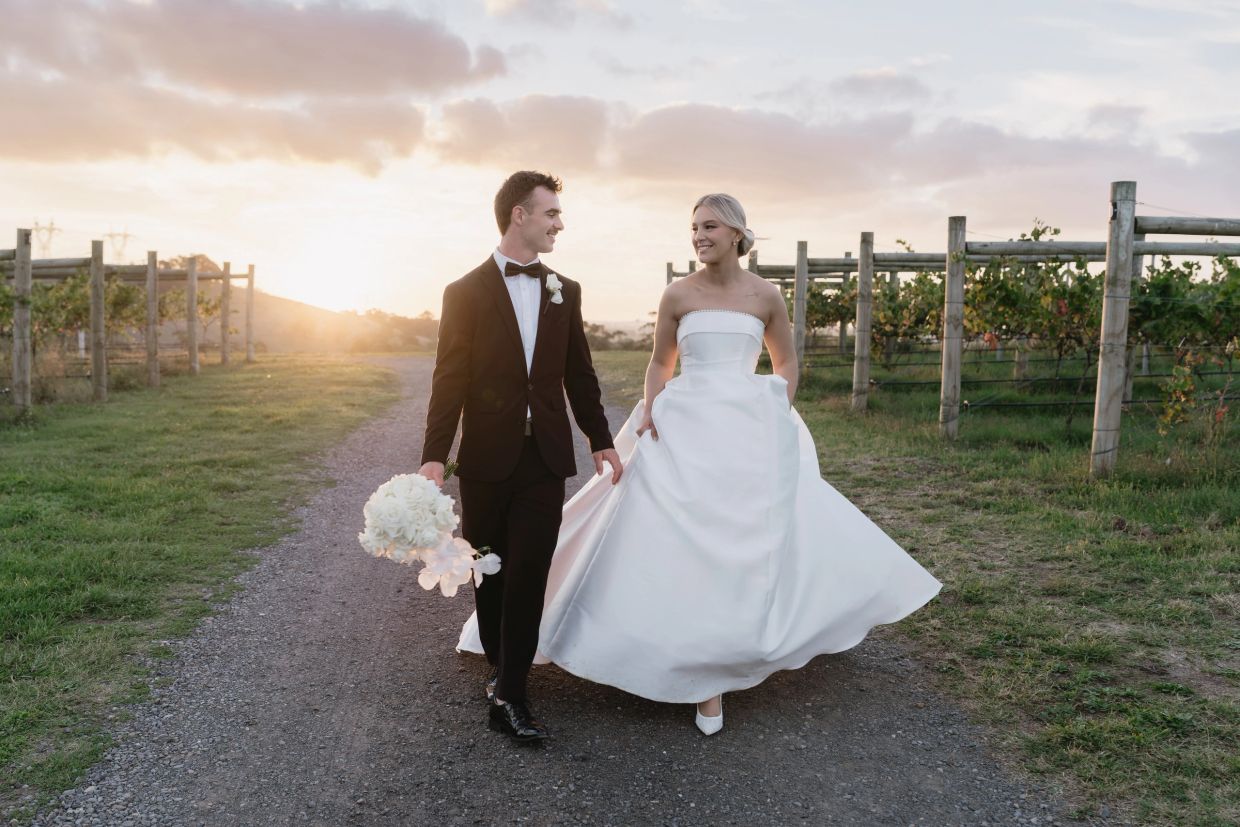 Bride and groom walking on a vineyard path at sunset.