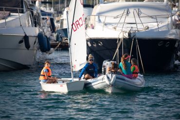 Children and adults on small boats near yachts in a marina.