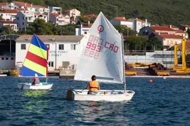 Two small sailboats with colorful sails on calm water near a coastal town.