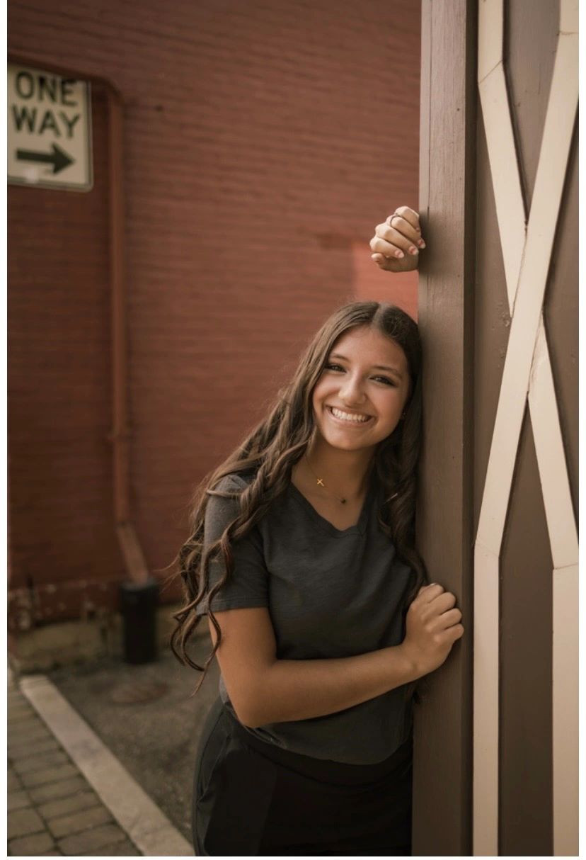 Smiling young woman leaning on a wooden post near a brick wall and one-way sign.