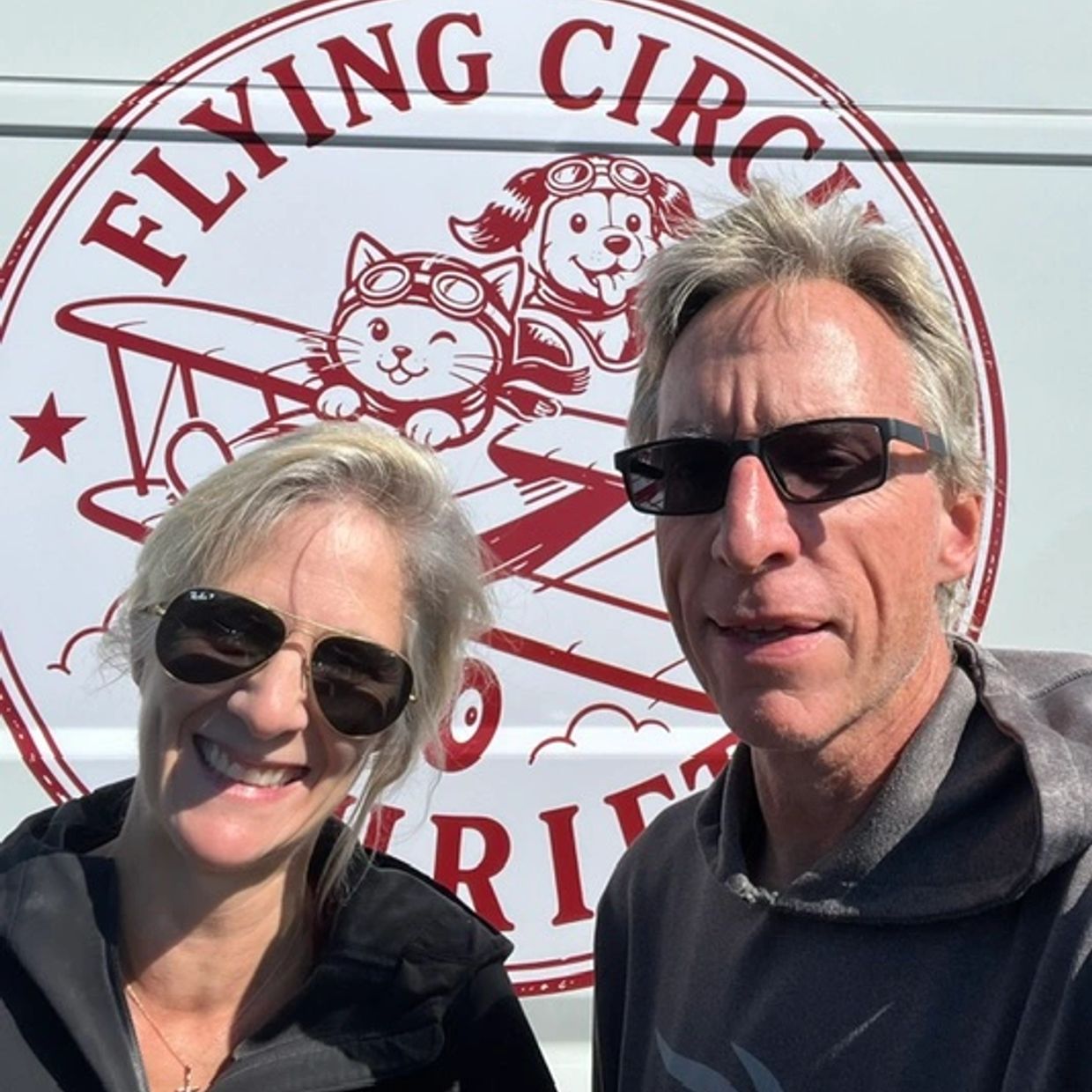 Two people wearing sunglasses smiling in front of a Flying Circus van.