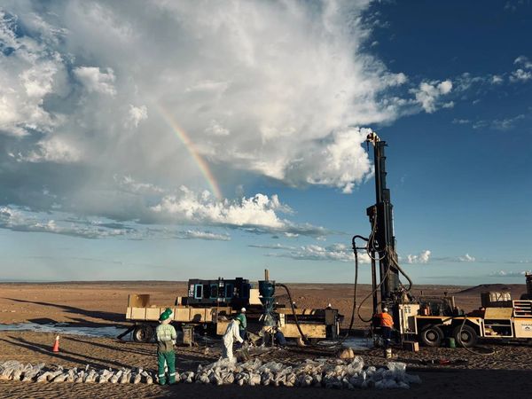 Workers operate drilling equipment in a desert with a rainbow overhead.