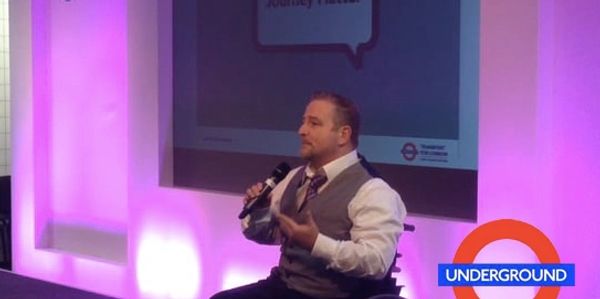 Ruben, in a wheelchair, is speaking to an audience with a microphone while wearing a white shirt.