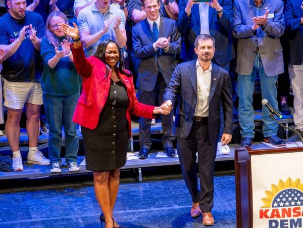Two political candidates hold hands and wave to an applauding crowd on stage.