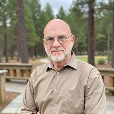 Man with a bald head and glasses, standing outdoors in a forested area.