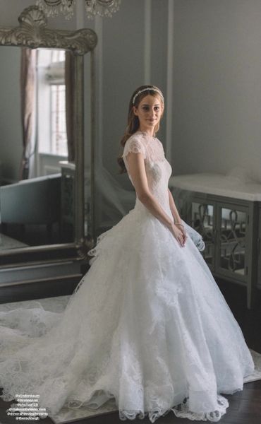 Bride in a lace white wedding gown posing indoors by a large mirror.
