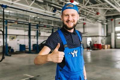 Smiling mechanic in blue overalls giving a thumbs up in a spacious workshop.