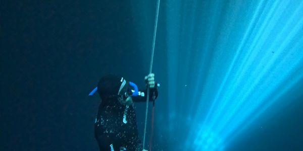 A scuba diver ascends underwater holding a rope with blue light beams.