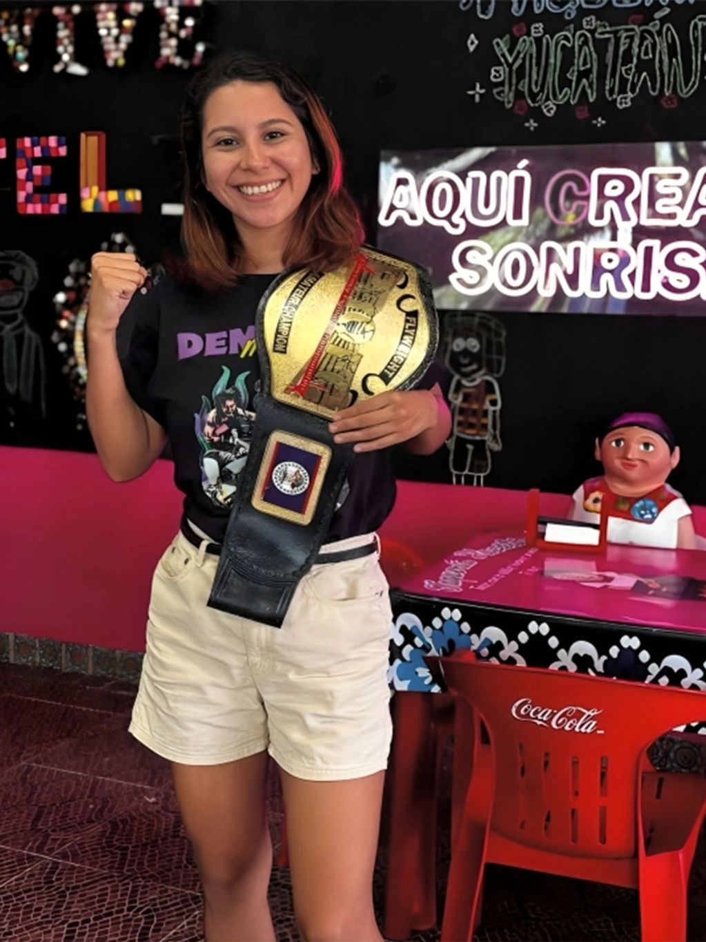 Smiling woman proudly holds a championship belt and poses with a fist raised.