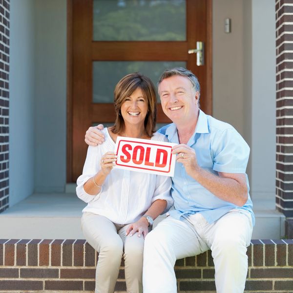 Happy mature couple sitting in front of a house holding a sold sign, Belair conveyancer