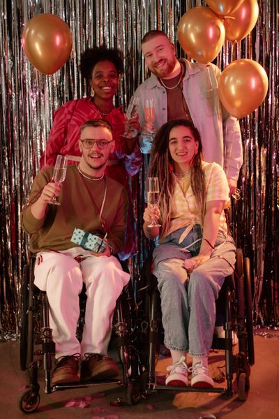 Four friends celebrating with champagne and balloons, two in wheelchairs.