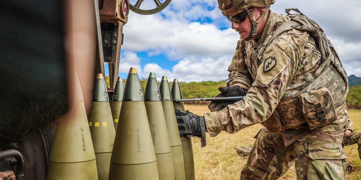 Soldier preparing large artillery shells outdoors under a partly cloudy sky.