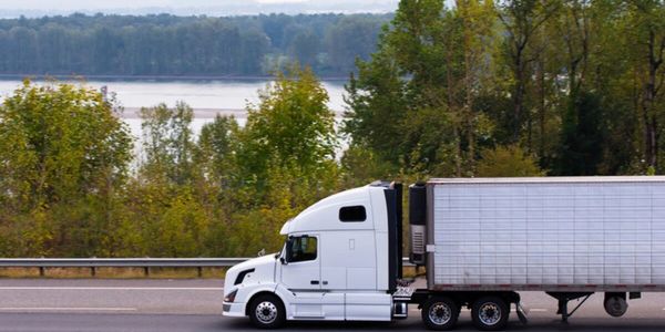 A white semi-truck driving on a highway with trees and a river in the background.