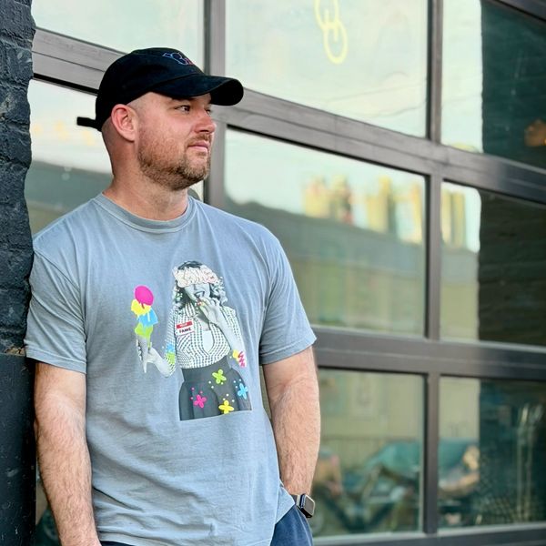 Man in a black cap and graphic t-shirt leaning against a wall.