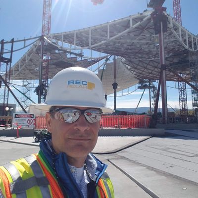 Construction worker wearing safety gear at a large site with cranes and a steel structure.