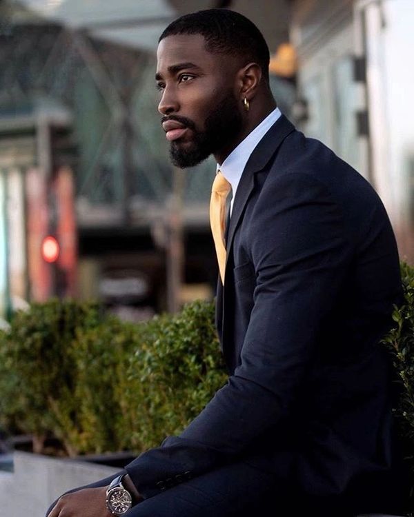 A well-dressed man in a navy suit with a yellow tie, sitting thoughtfully outdoors.