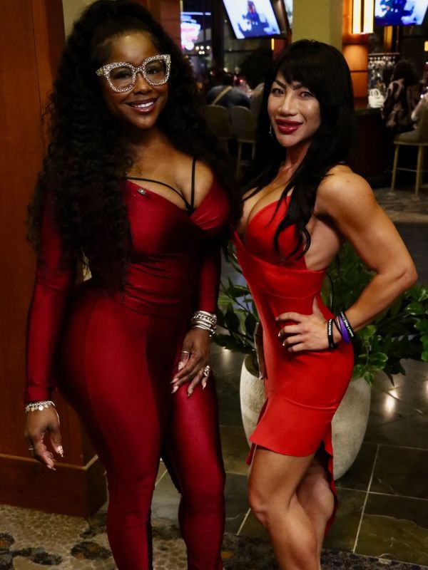 Two women dressed in elegant red outfits pose indoors with smiles.