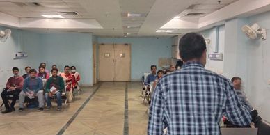 A man addressing a group seated in a room with blue walls and ceiling fans.