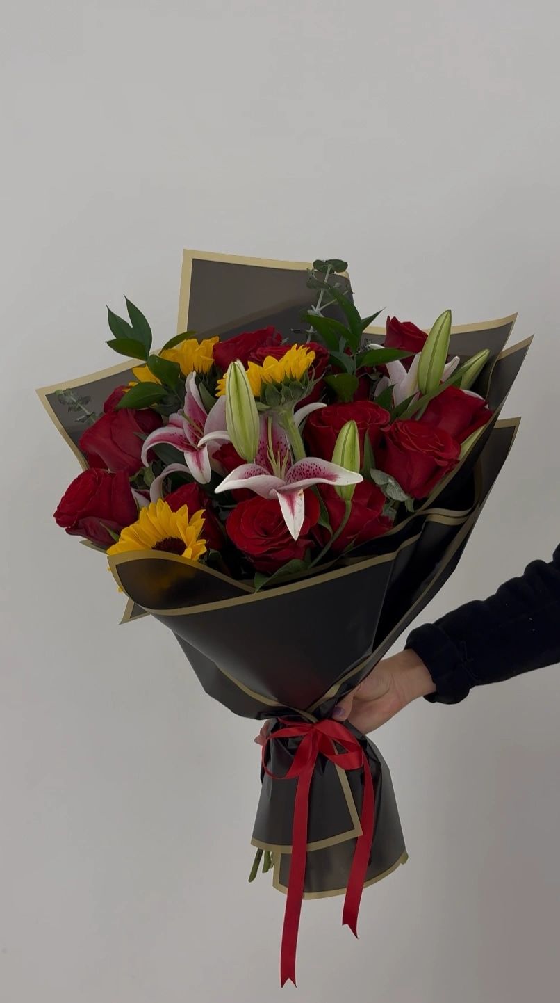A vibrant bouquet of red roses, sunflowers, and lilies wrapped in black paper with a red ribbon.