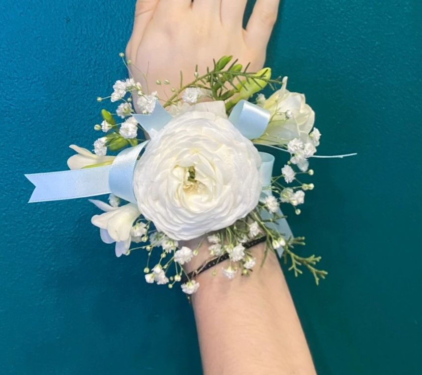 White floral corsage with blue ribbon on wrist against teal background.