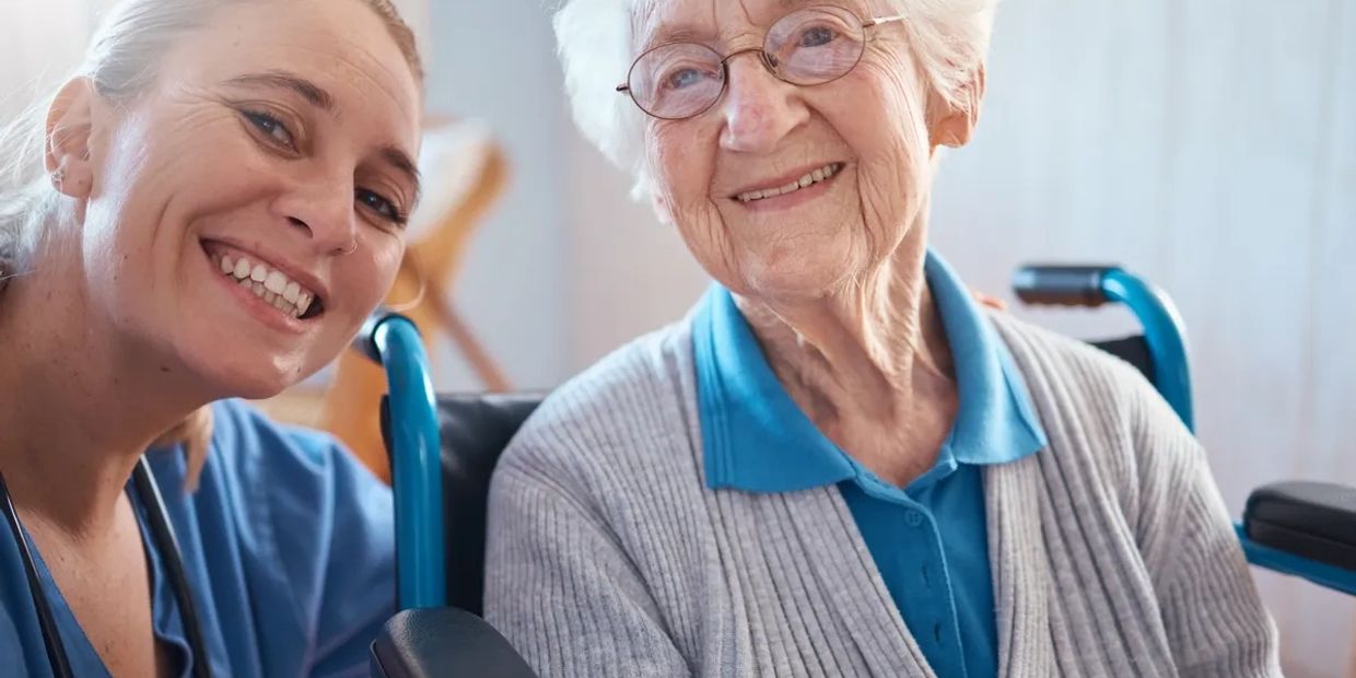 Smiling elderly woman with caregiver in blue scrubs, sharing a happy moment.