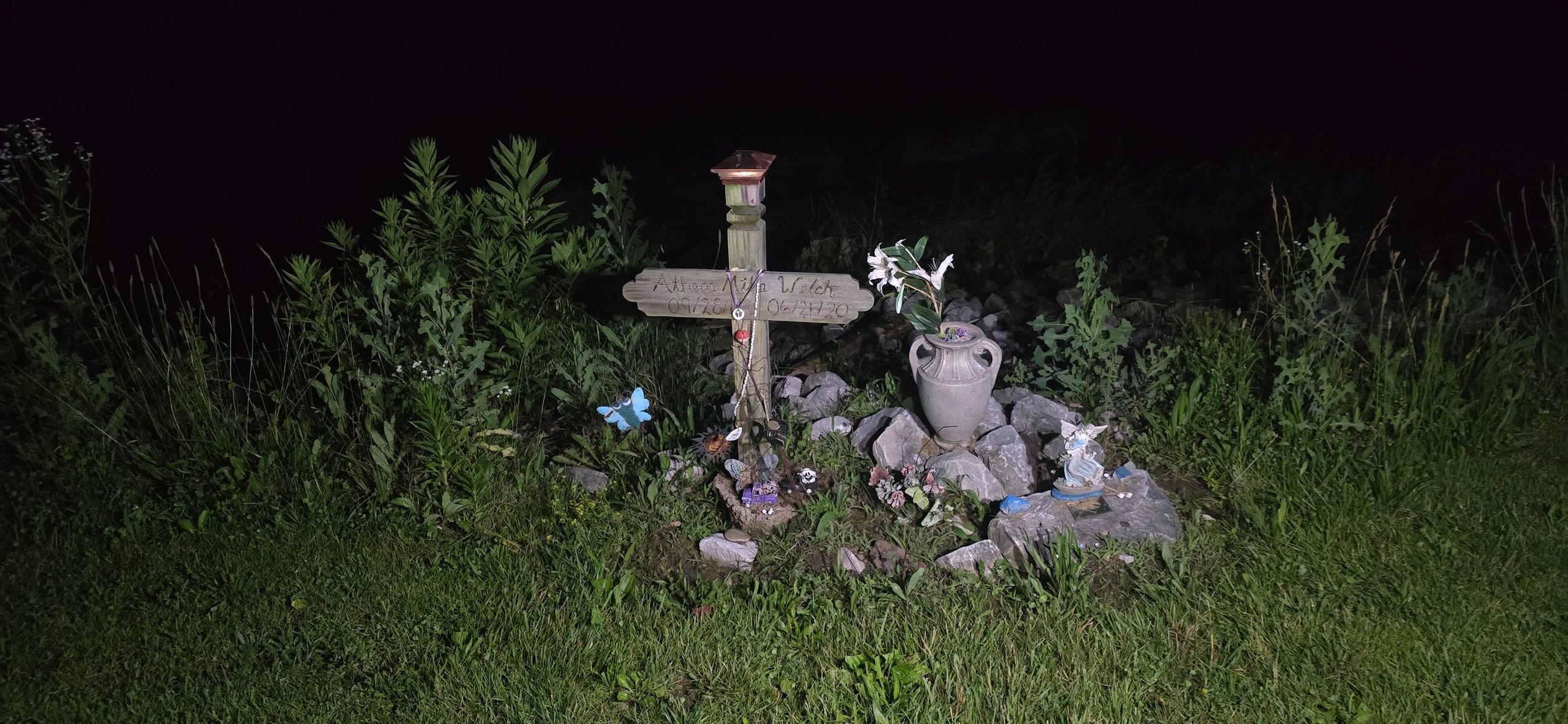 A nighttime memorial cross with flowers and decorations in a grassy area.