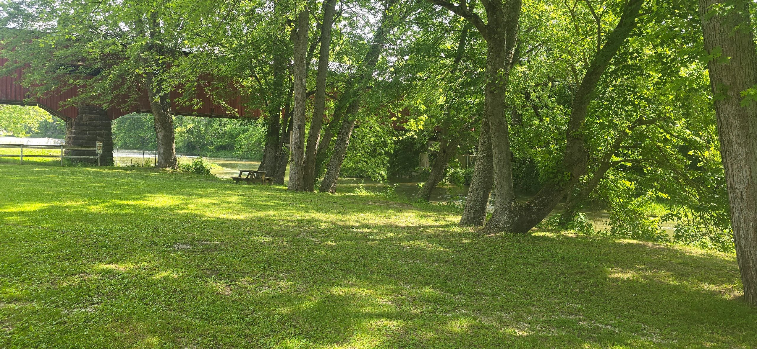 A serene park with green grass, tall trees, and a red covered bridge over a river.