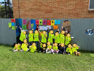 Group of children in matching green shirts posing outdoors in front of colorful artwork on a fence.