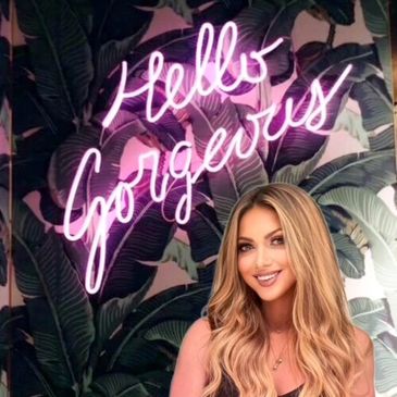 Smiling woman with long blonde hair in front of a neon "Hello Gorgeous" sign.