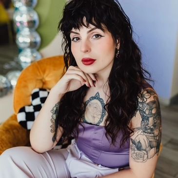 Young woman with dark curly hair and tattoos sits on an orange chair.