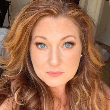 A close-up portrait of a woman with curly red hair and blue eyes.