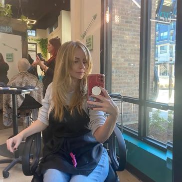 Woman in a salon taking a selfie with a phone, hairdresser working in background.