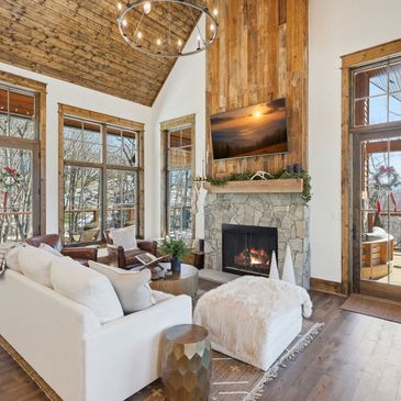 Cozy rustic living room with large windows, stone fireplace, and wood accents.