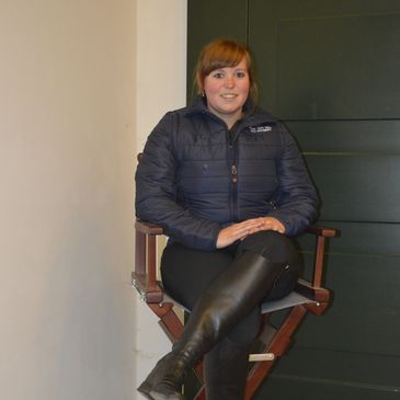 Vrouw zittend op een hoge stoel, gekleed in een donker jasje en laarzen.