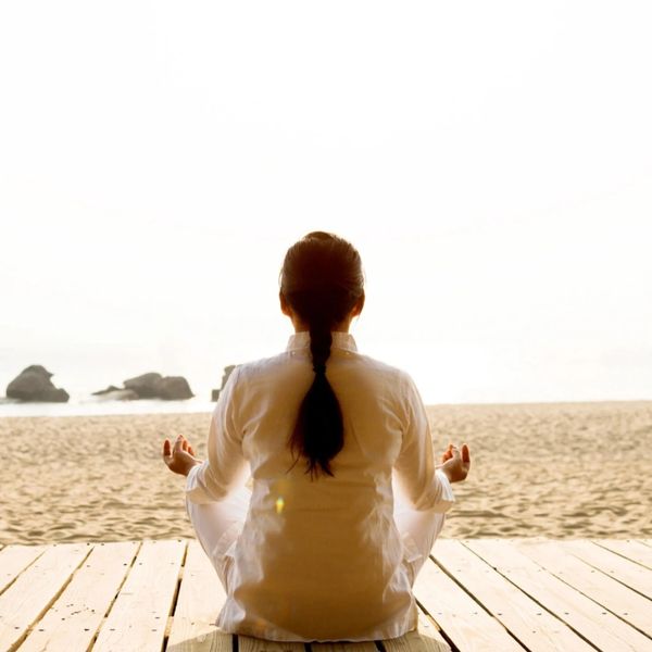 Person meditating on a wooden deck facing the beach at sunrise.